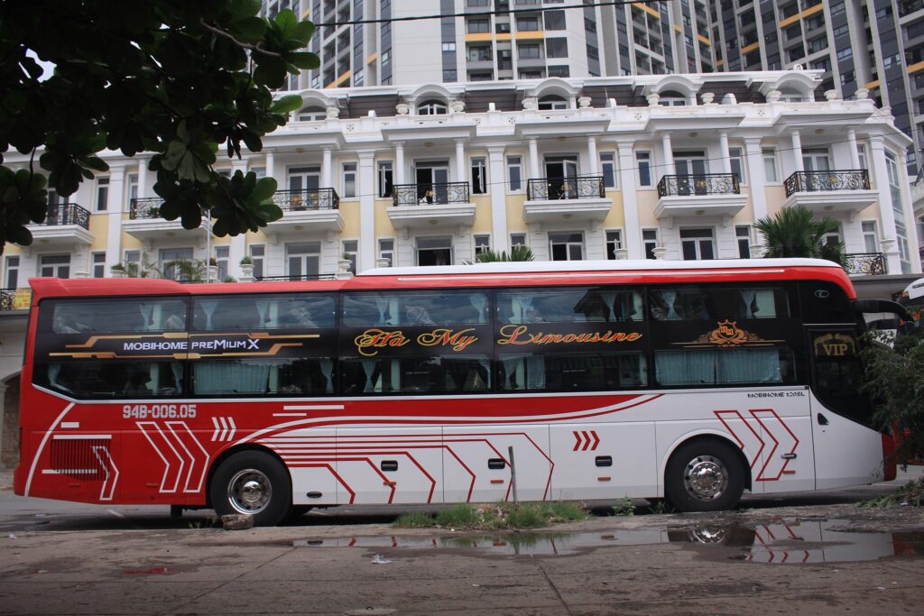một oto màu đỏ sọc trắng phái sau có cành cây và tòa nhà màu trắngthuê xe du lịch Ninh Thuận