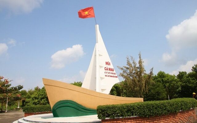 Có 1 chiếc thuyền mô hình, xung quanh là những hàng cây bên trên là bầu trời xanh và Cờ Tổ Quốc Việt Nam